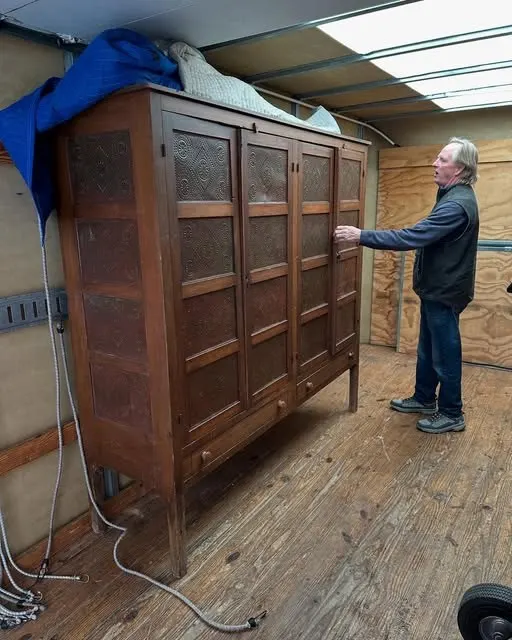 A tall wooden cupboard with punched-tin panels being checked over inside a moving truck